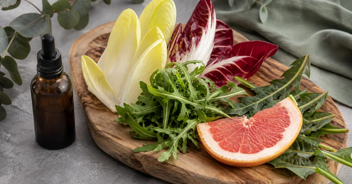 Holzbrett mit bitteren Zutaten: Chicorée, Radicchio, Rucola, Löwenzahnblätter und eine Grapefruitscheibe, daneben ein Fläschchen Bittertropfen bei natürlichem Licht.